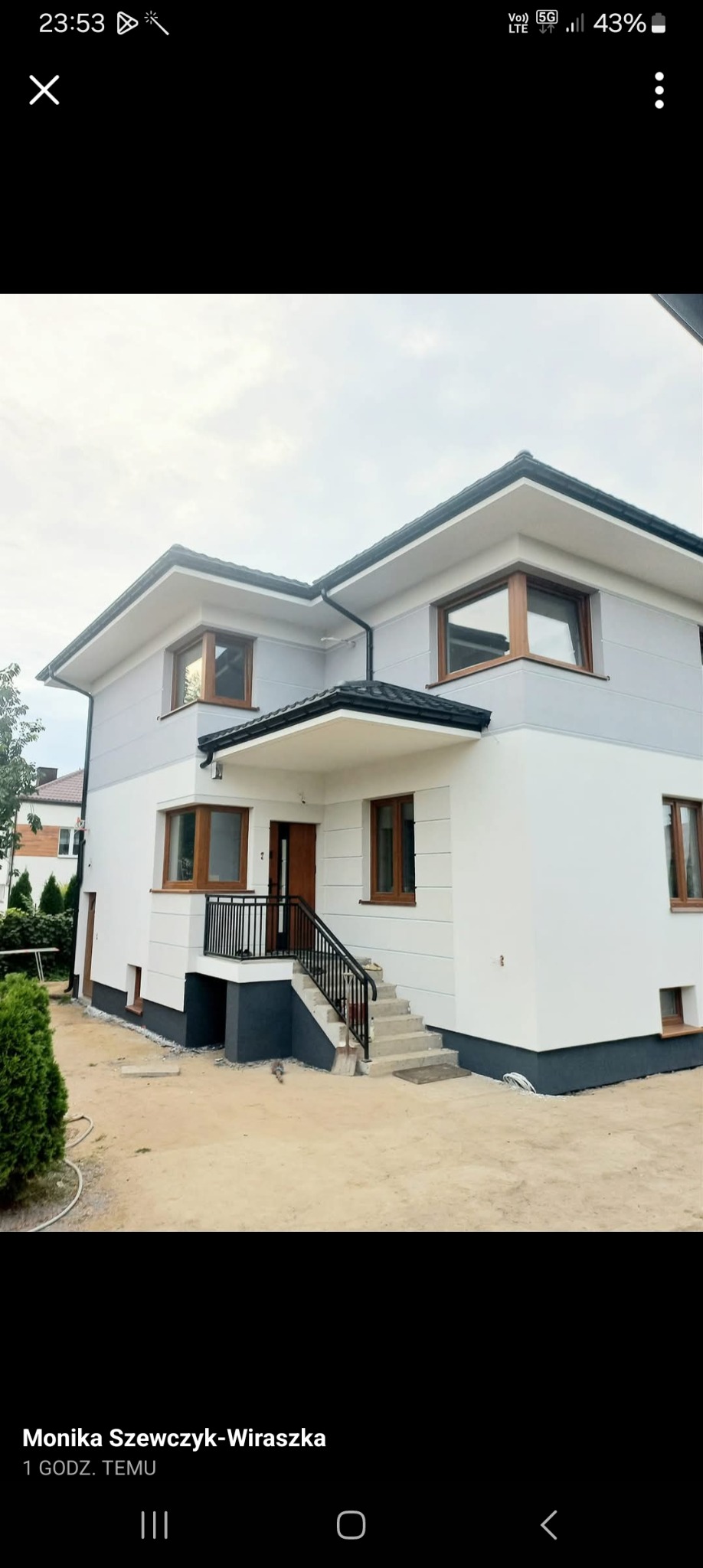 Dwukondygnacyjny dom z jasną elewacją, ciemnymi oknami i schodami wejściowymi. Widoczne fragmenty ogrodu. Ujęcie z poziomu gruntu.