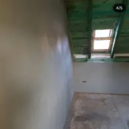 Gładka, jasna ściana po nałożeniu tynku maszynowego, widoczne drewniane elementy konstrukcji dachu i okno połaciowe w surowym stanie.