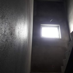 Patryk Łukowski - Ściana klatki schodowej po nałożeniu szarego tynku maszynowego, widoczne schody i okno z intensywnym światłem dziennym.