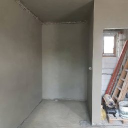 Patryk Łukowski - Świeżo nałożona, gładka warstwa szarego tynku na ścianach i podłodze pomieszczenia w trakcie remontu. Widoczne okno w stanie surowym, drabina i nagrzewnica obok.