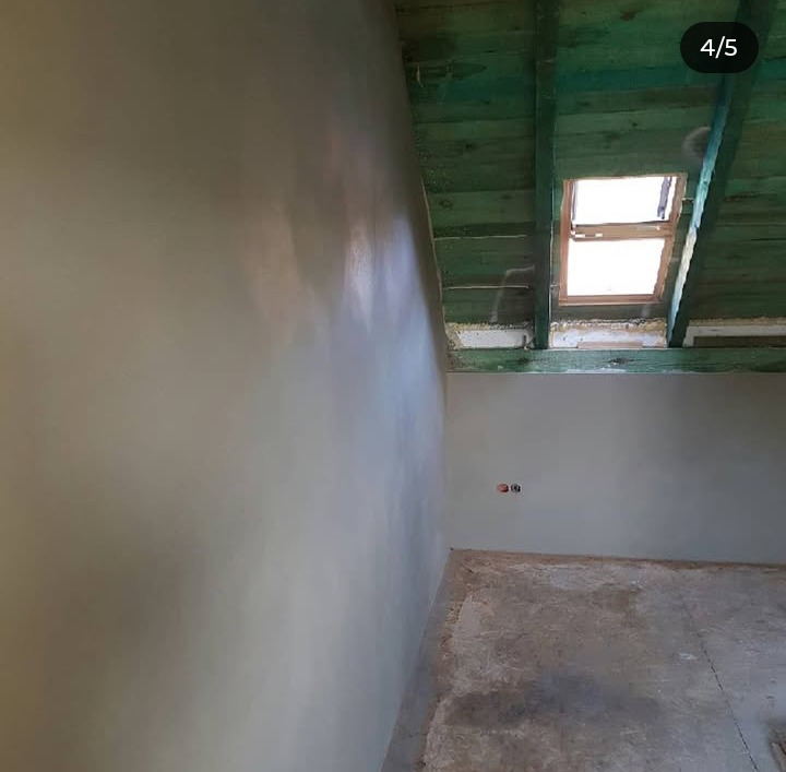 Gładka, jasna ściana po nałożeniu tynku maszynowego, widoczne drewniane elementy konstrukcji dachu i okno połaciowe w surowym stanie.