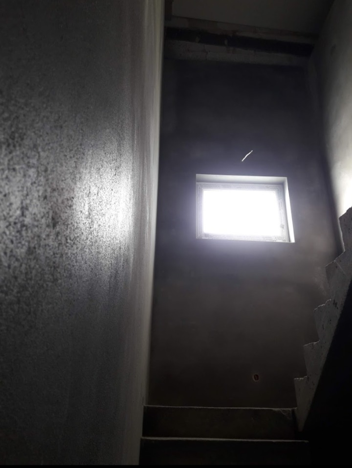 Ściana klatki schodowej po nałożeniu szarego tynku maszynowego, widoczne schody i okno z intensywnym światłem dziennym.