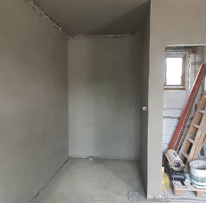 Świeżo nałożona, gładka warstwa szarego tynku na ścianach i podłodze pomieszczenia w trakcie remontu. Widoczne okno w stanie surowym, drabina i nagrzewnica obok.