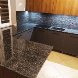 ZAKŁAD OBRÓBKI KAMIENIA NATURALNEGO RSGranit ROMANA JANKOWSKA - Kuchnia z ciemnym blatem z kamienia kompozytowego o drobnej fakturze, szare szafki z poziomymi uchwytami, drewniane szafki wiszące i podświetlenie LED nad blatem.