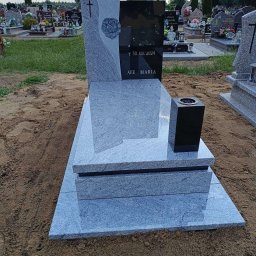 ZAKŁAD OBRÓBKI KAMIENIA NATURALNEGO RSGranit ROMANA JANKOWSKA - Granitowy nagrobek z wygrawerowanym krzyżem i różą, z czarną tablicą inskrypcyjną i wazonem na cmentarzu.