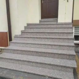 Zewnętrzne schody wejściowe z szarego, nakrapianego kamienia prowadzące do brązowych drzwi wejściowych.