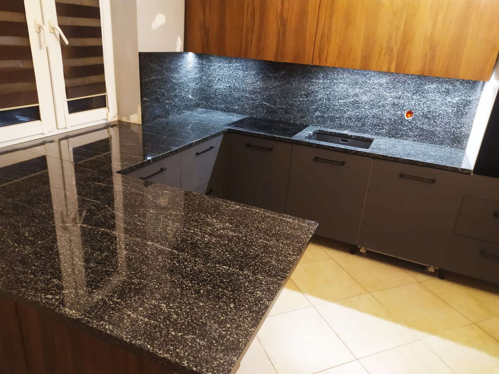 Kuchnia z ciemnym blatem z kamienia kompozytowego o drobnej fakturze, szare szafki z poziomymi uchwytami, drewniane szafki wiszące i podświetlenie LED nad blatem.