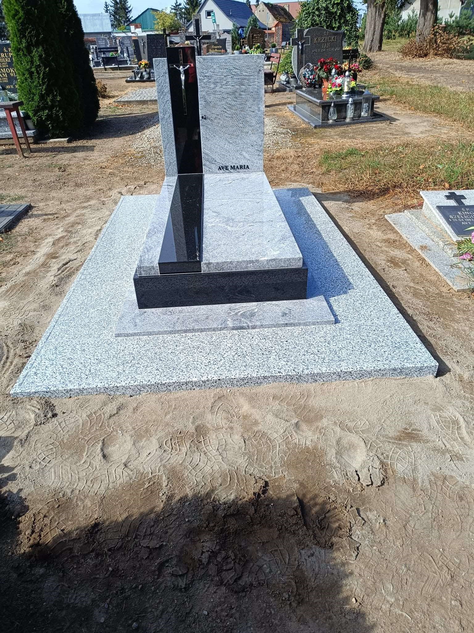Granitowy pomnik nagrobny z czarnym akcentem i napisem 'Ave Maria' na cmentarzu, widoczne ślady stóp na piasku wokół.