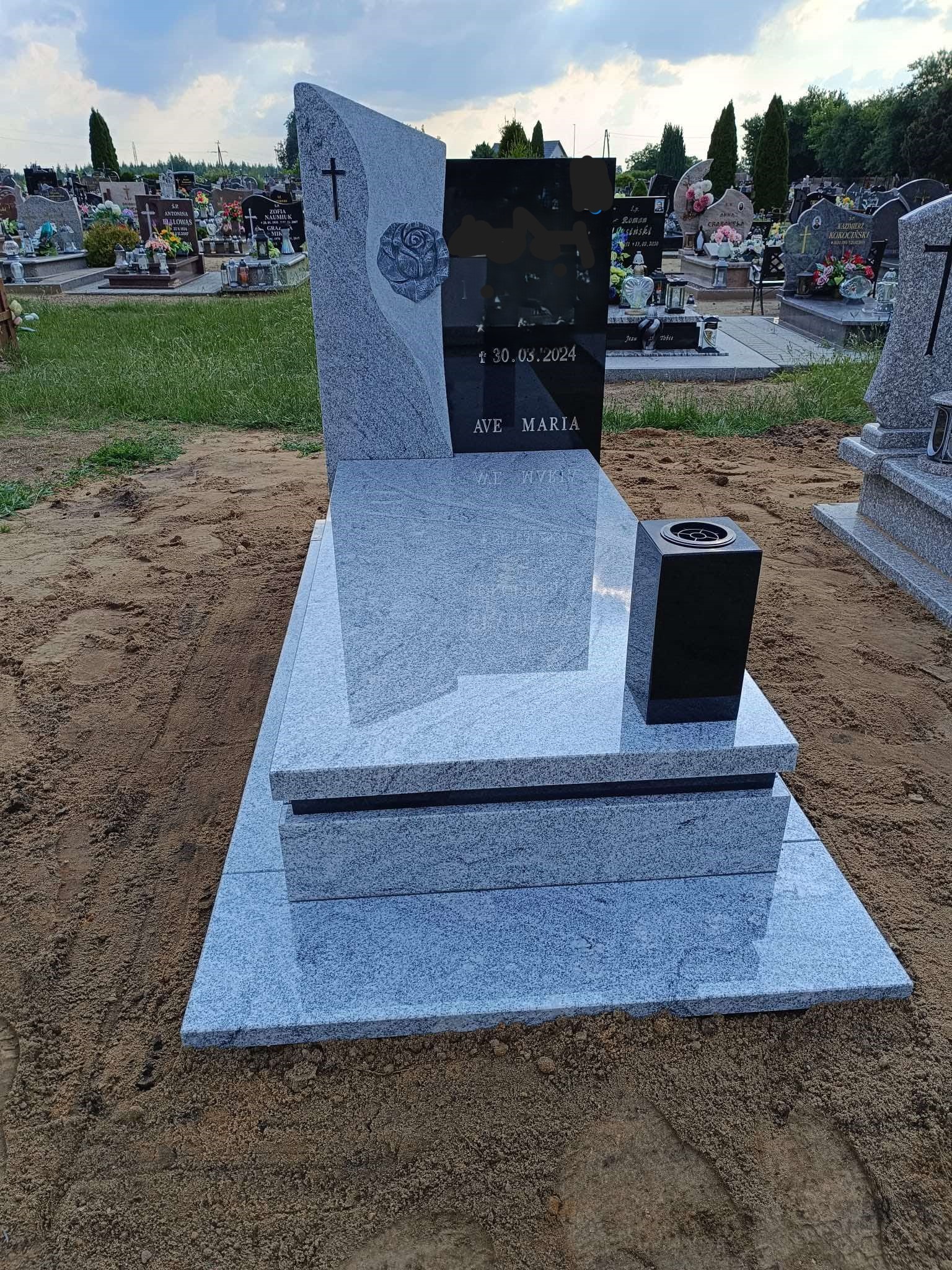 Granitowy nagrobek z wygrawerowanym krzyżem i różą, z czarną tablicą inskrypcyjną i wazonem na cmentarzu.