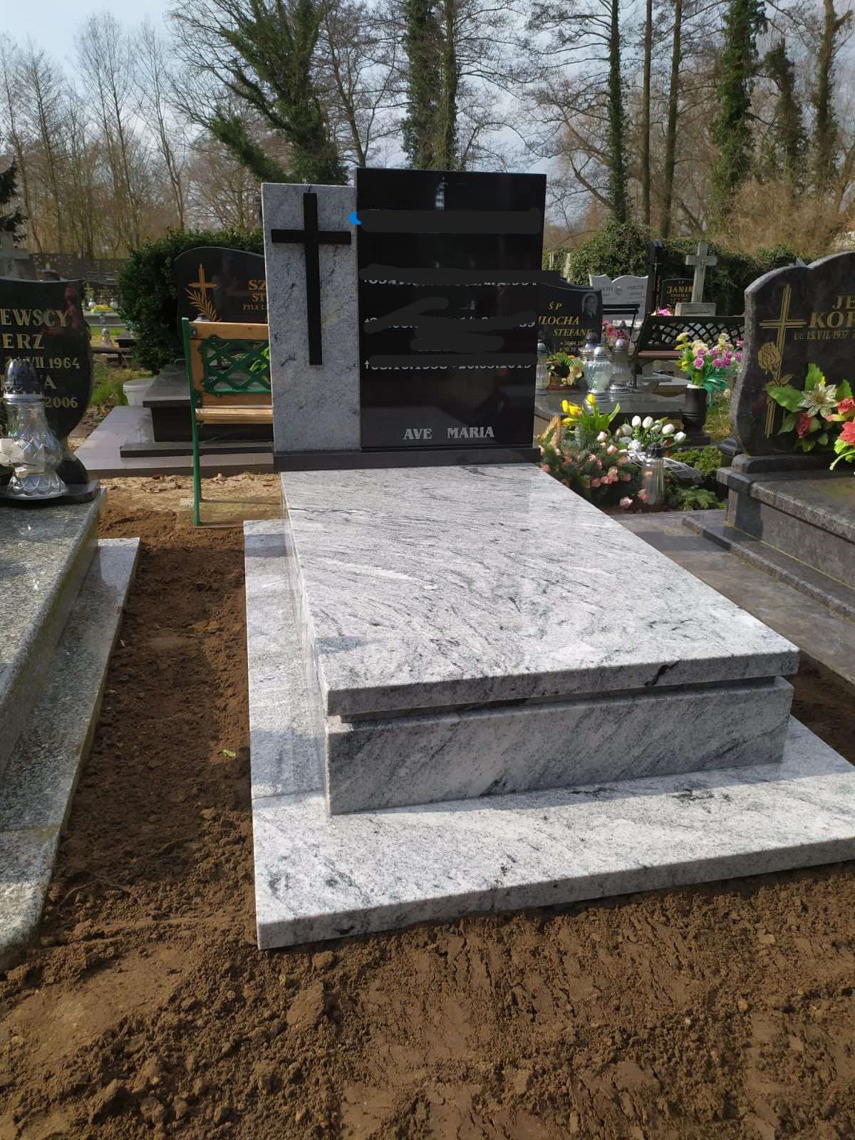 Jasnoszary, granitowy nagrobek z czarną tablicą i krzyżem na cmentarzu, otoczony ziemią.