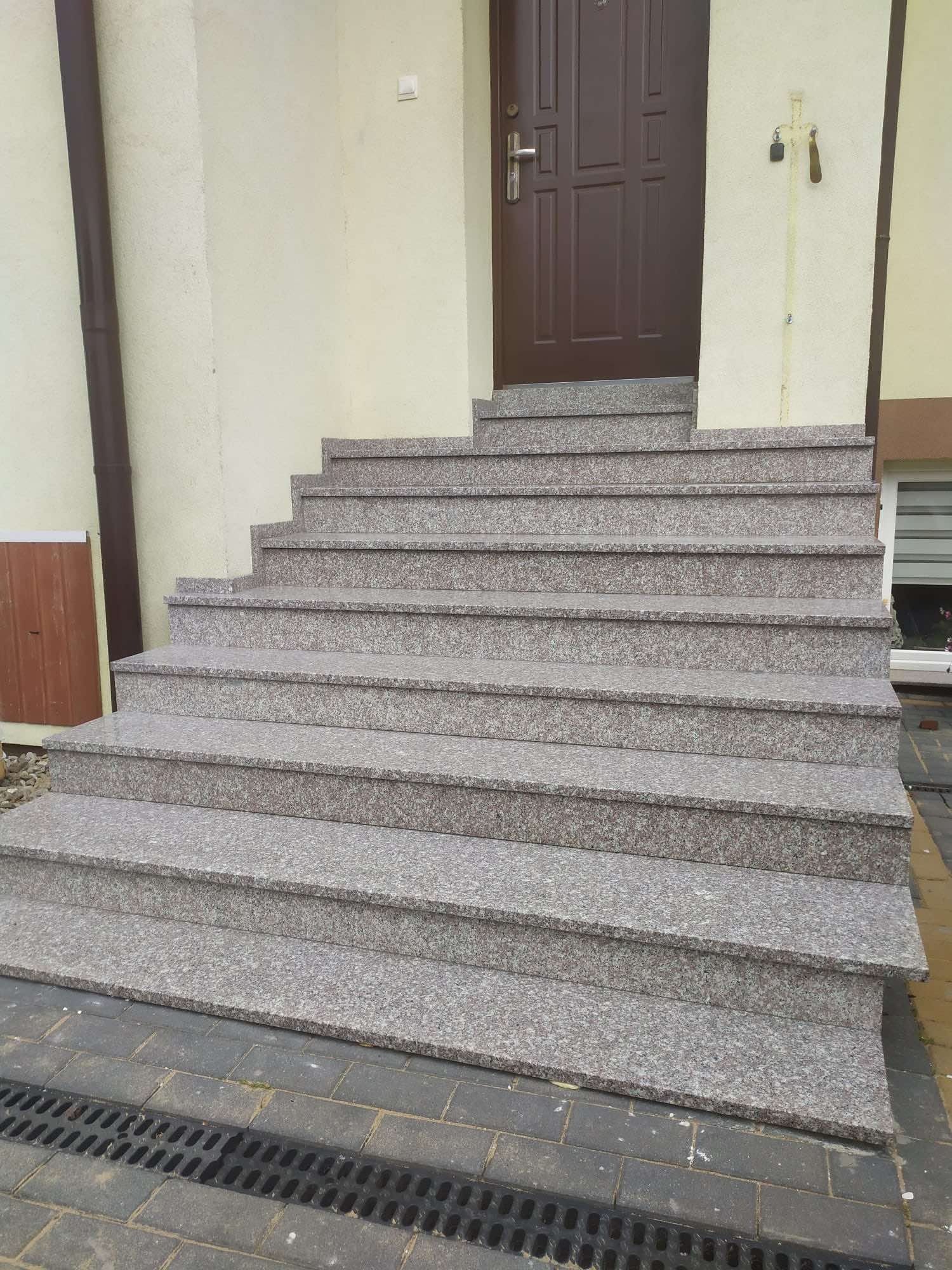 Zewnętrzne schody wejściowe z szarego, nakrapianego kamienia prowadzące do brązowych drzwi wejściowych.