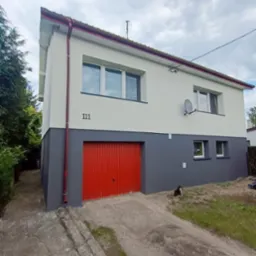 Dwukondygnacyjny dom z szarą dolną i białą górną częścią elewacji, czerwonymi drzwiami garażowymi i numerem 111. Na trawniku siedzi czarno-biały kot.