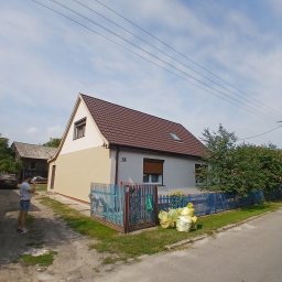 Gentle Investments - Dom jednorodzinny po odświeżeniu elewacji w odcieniach beżu i bieli, z brązowym dachem. Widoczne worki z odpadami po pracach. Niebieskie ogrodzenie i osoba w tle.