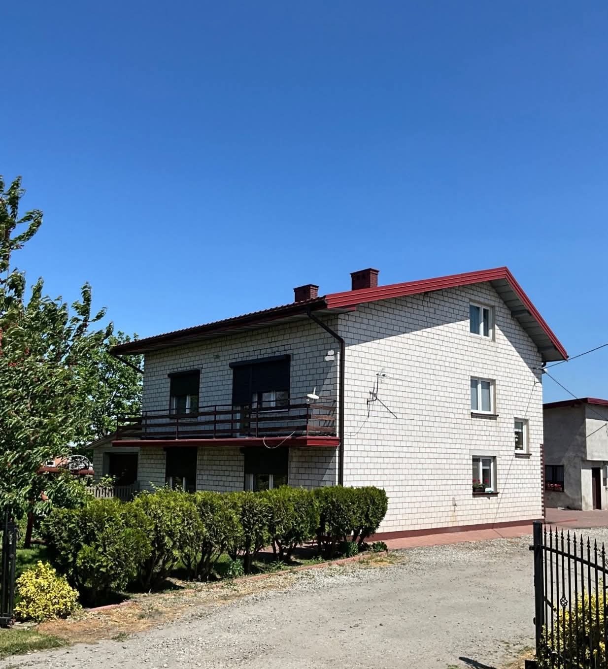 Dwupiętrowy dom z białej cegły z czerwonym dachem i balkonem, otoczony zielenią. Widoczne krzewy i drzewo. Błękitne niebo w tle. Dom po renowacji elewacji.