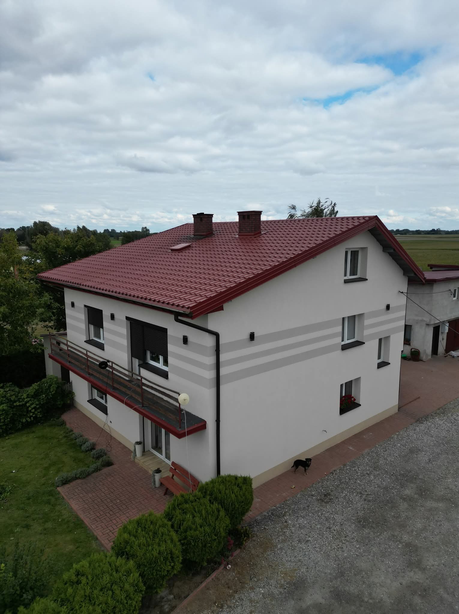 Dwupiętrowy dom z czerwonym dachem, białymi ścianami z szarymi pasami i balkonem. Widoczny ogród, kostka brukowa i pies. Ujęcie z góry, pochmurne niebo.