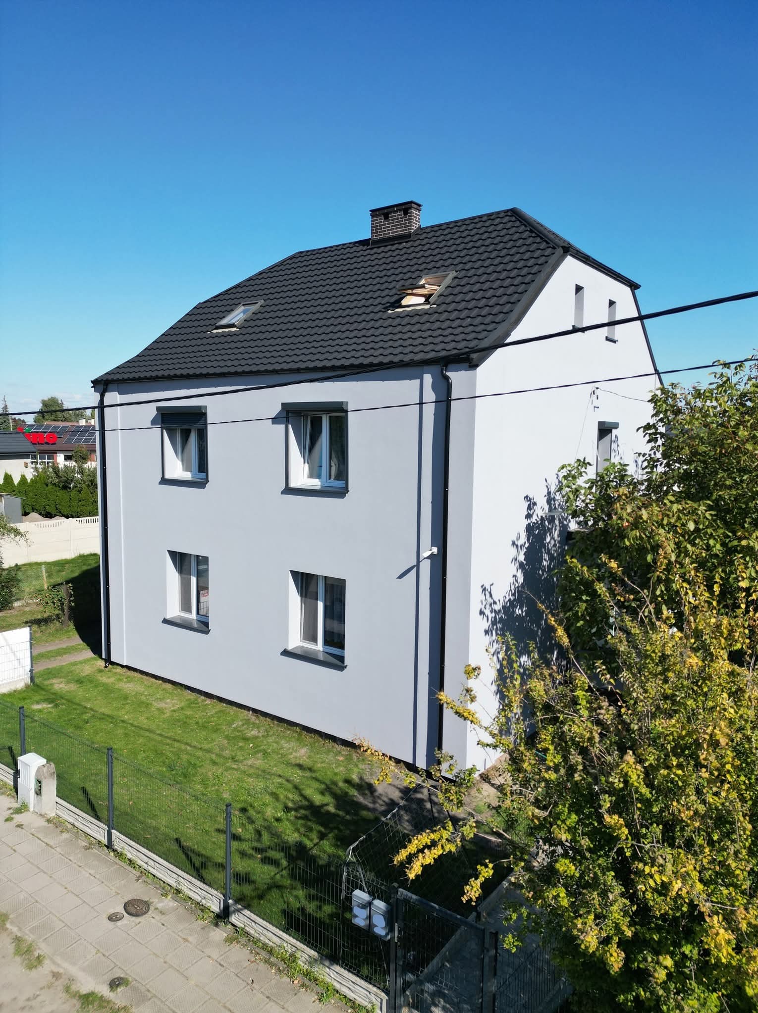 Dwukondygnacyjny dom z szarą elewacją i ciemnym dachem, widziany z góry. Na dachu okna połaciowe. W tle zieleń i błękitne niebo.