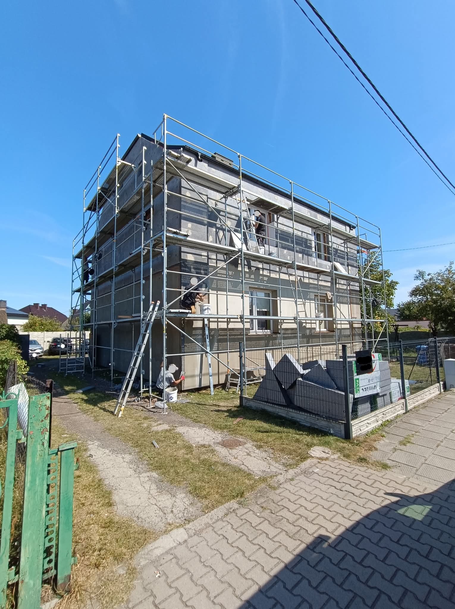 Dom w rusztowaniach podczas prac elewacyjnych. Widoczni pracownicy, materiały izolacyjne i przygotowane podłoże. Słoneczny dzień, błękitne niebo.