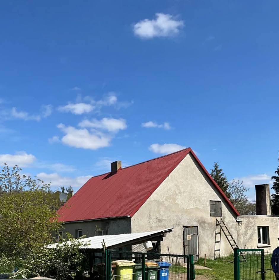 Dom z czerwoną blachodachówką przed ociepleniem, z widoczną drabiną opartą o ścianę. W tle błękitne niebo z chmurami.