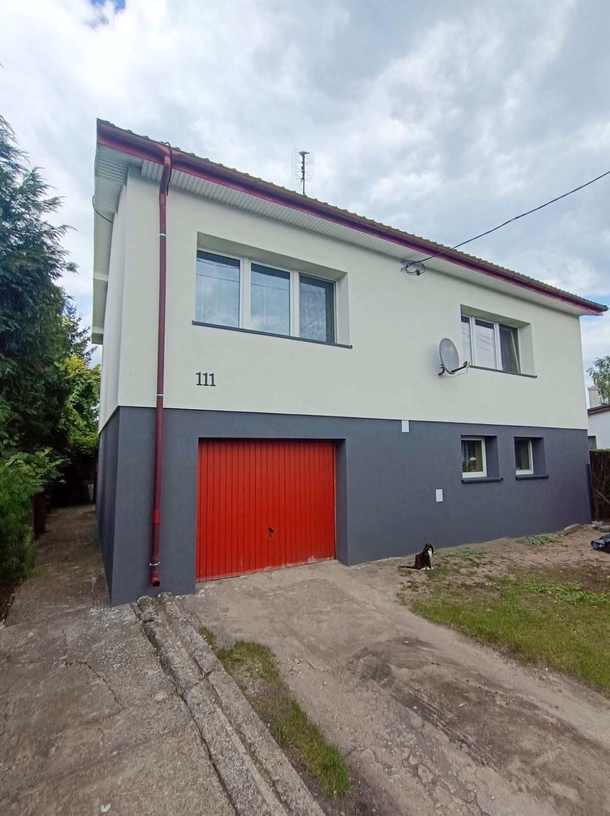 Dwukondygnacyjny dom z szarą dolną i białą górną częścią elewacji, czerwonymi drzwiami garażowymi i numerem 111. Na trawniku siedzi czarno-biały kot.