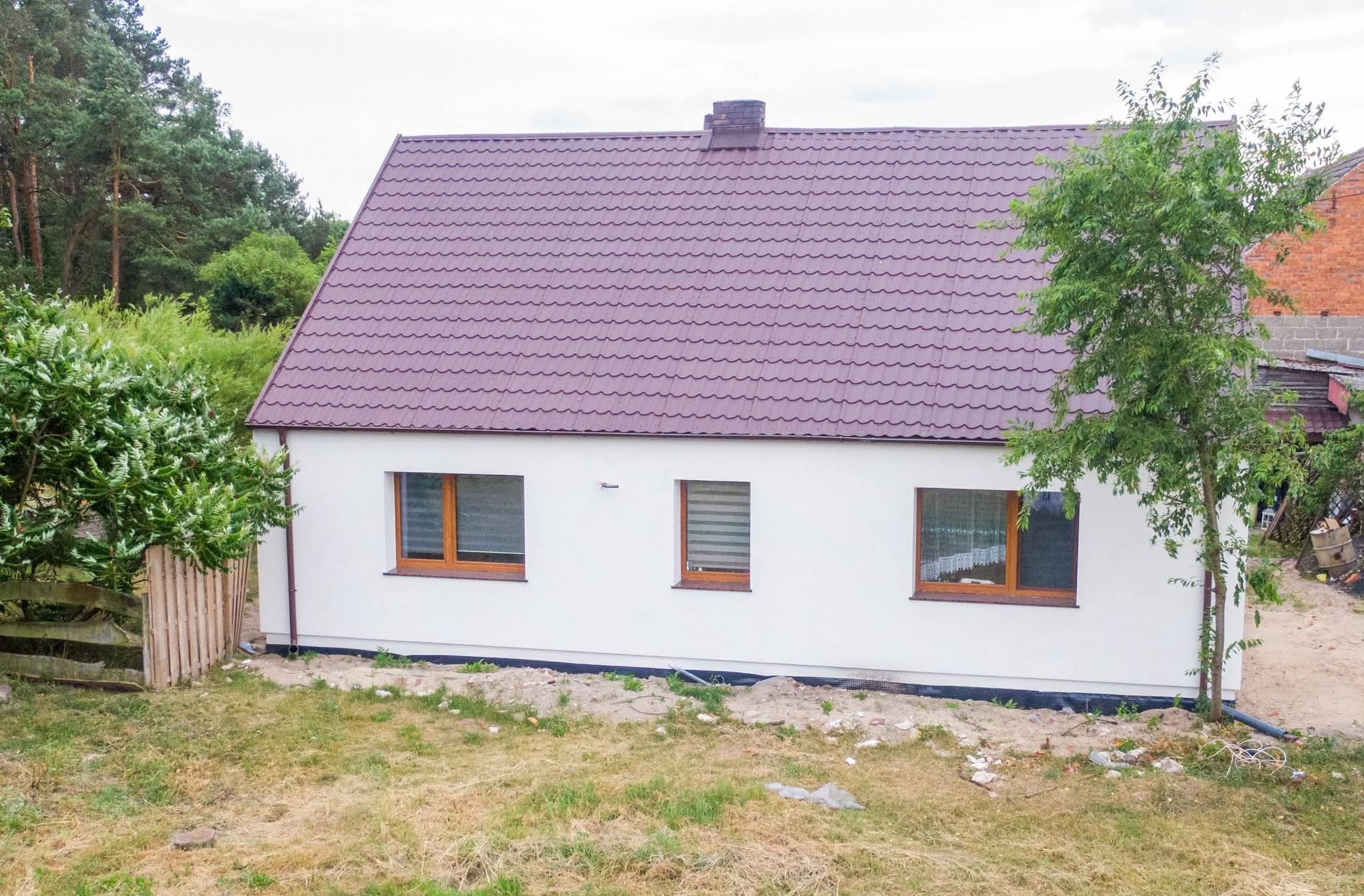 Biały dom z brązowym dachem z blachy trapezowej, widok z boku, otoczony zielenią. Widoczne trzy okna z drewnianą ramą i fragment drewnianego płotu.