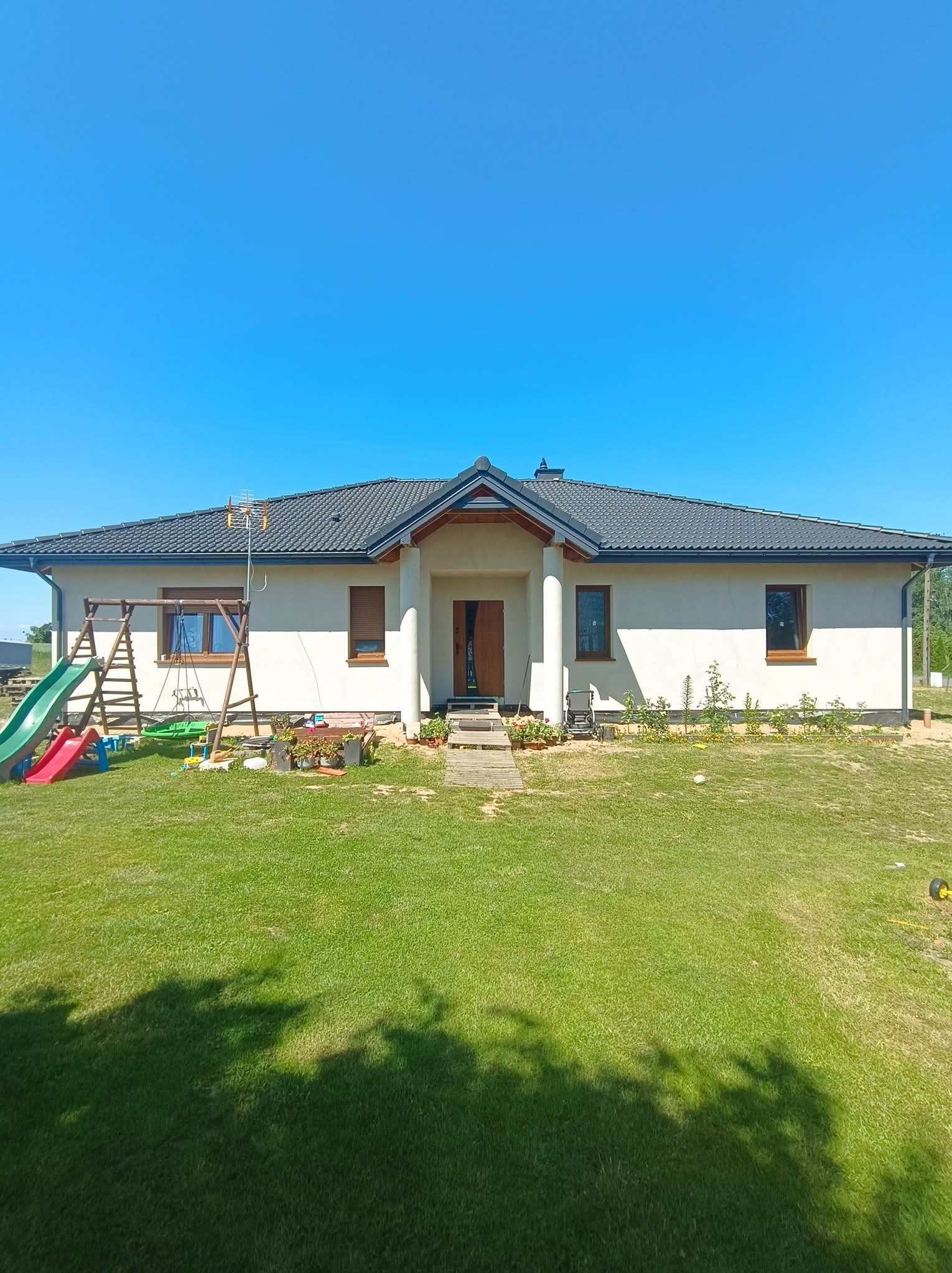 Jasny dom jednorodzinny z ciemnym dachem i drewnianymi oknami, otoczony zielonym trawnikiem i placem zabaw. Widok z przodu w słoneczny dzień.