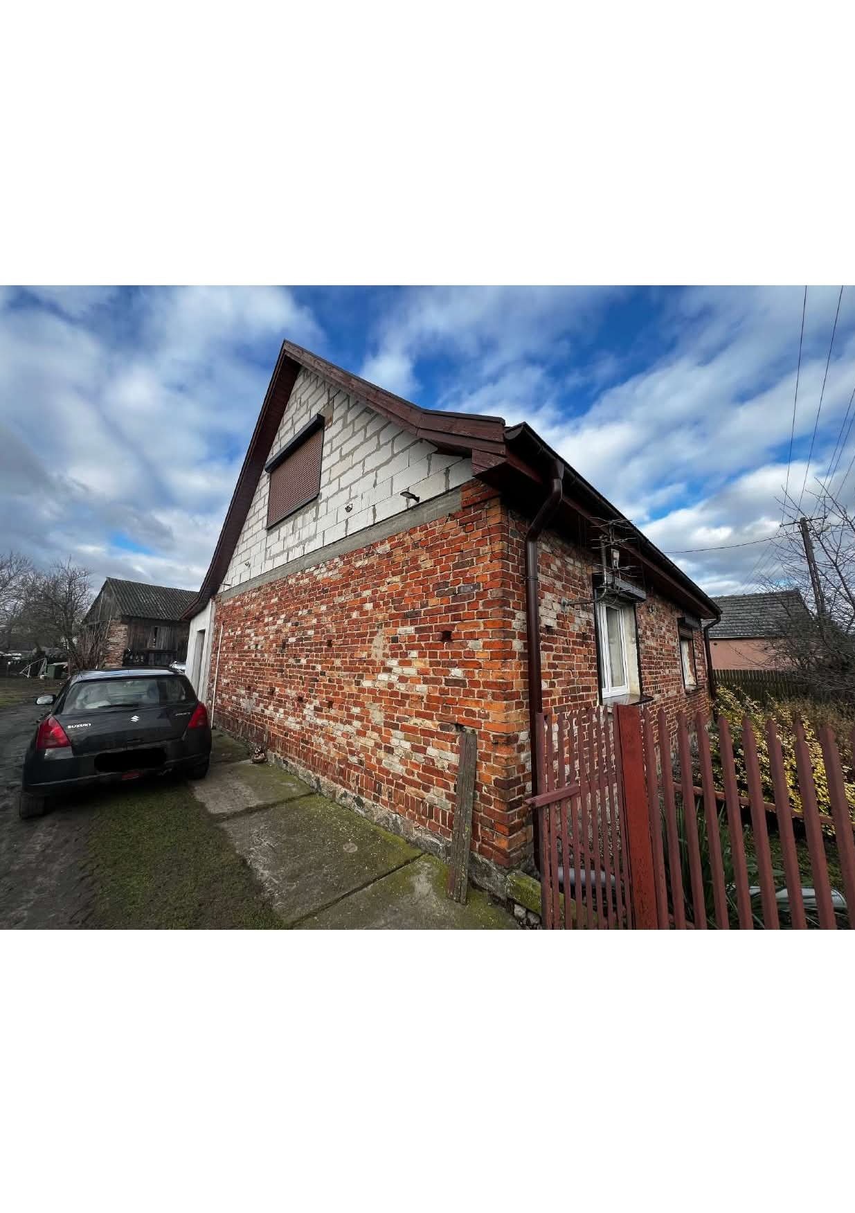 Dwukondygnacyjny dom z czerwonej cegły i białych bloczków, widoczny z boku, z zaparkowanym czarnym samochodem. Czerwone ogrodzenie i błękitne niebo w tle.