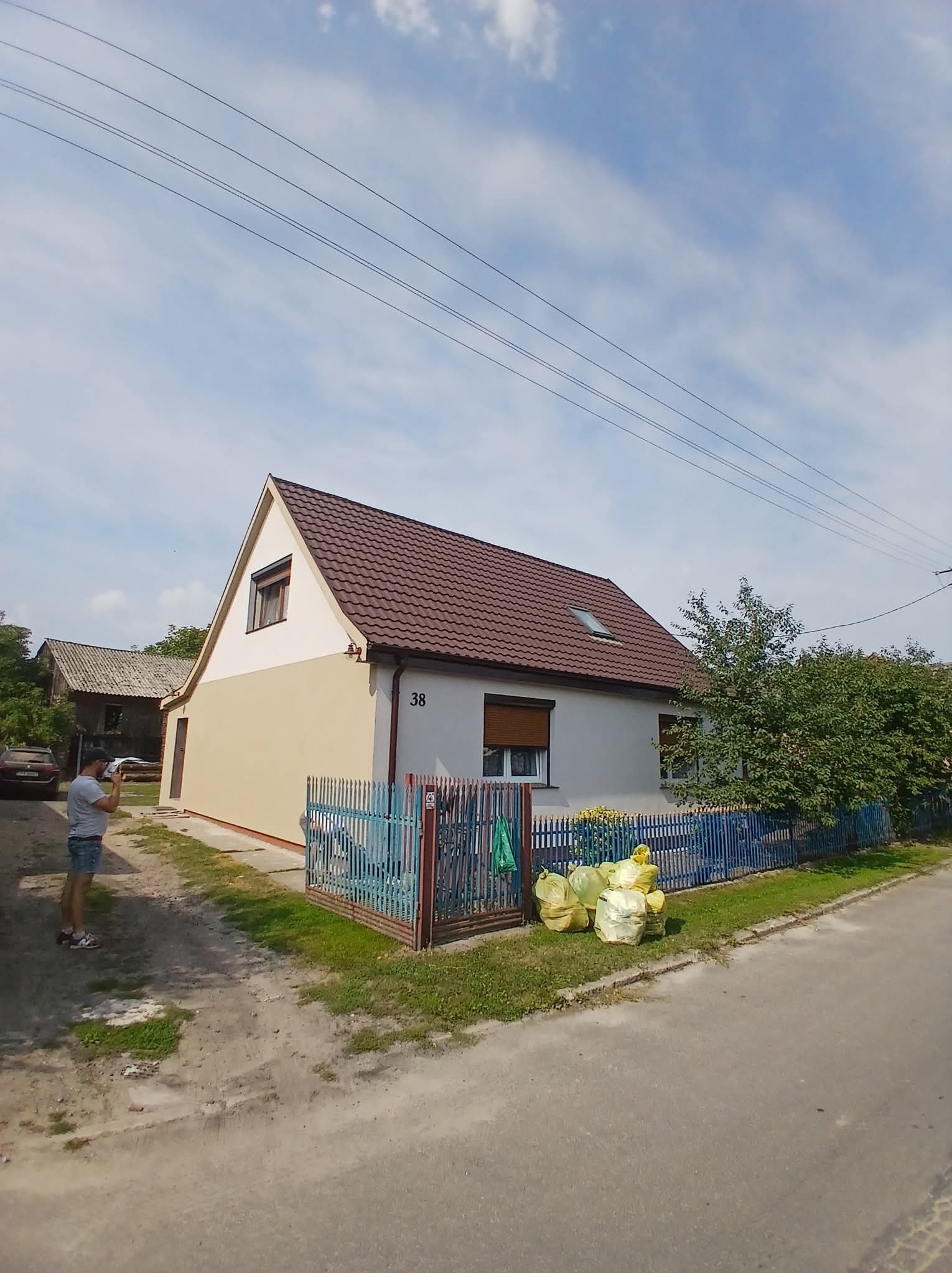 Dom jednorodzinny po odświeżeniu elewacji w odcieniach beżu i bieli, z brązowym dachem. Widoczne worki z odpadami po pracach. Niebieskie ogrodzenie i osoba w tle.