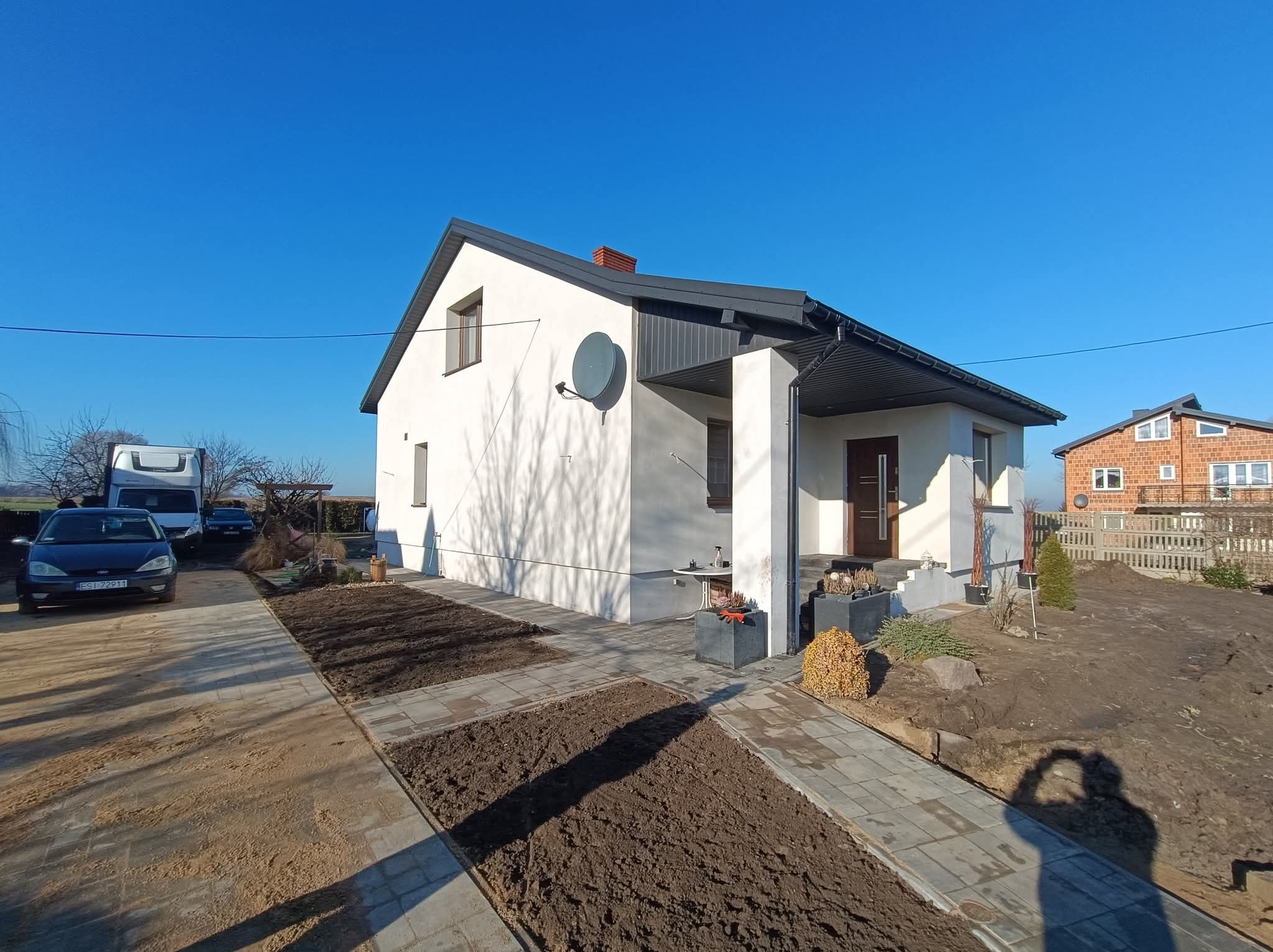 Biały dom jednorodzinny z szarą dachówką i elewacją po renowacji, widok z zewnątrz w słoneczny dzień. Uporządkowany teren wokół domu, świeżo przygotowane rabaty.