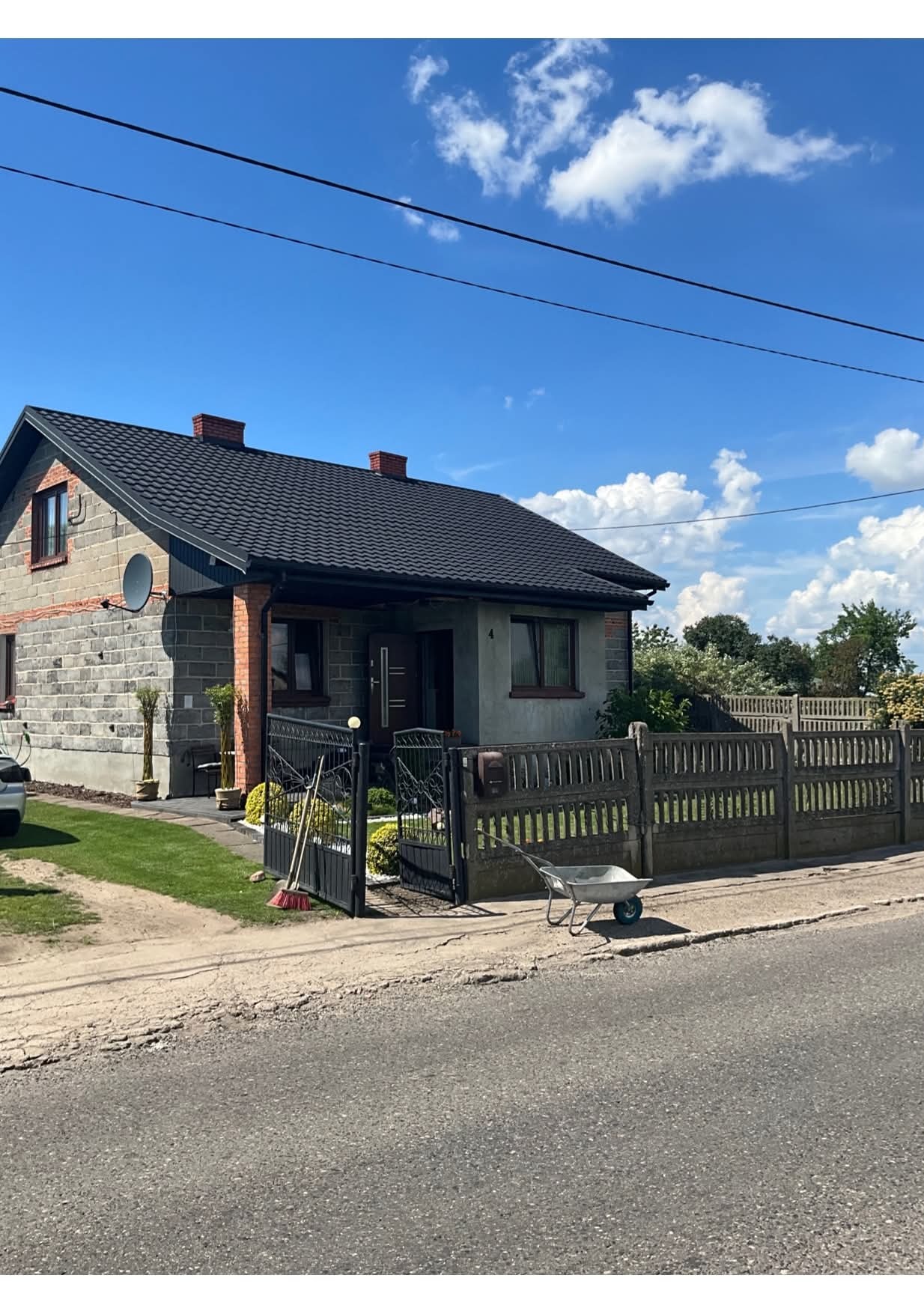 Dom jednorodzinny w Kaliszu z szarą elewacją z cegły i tynkowanym fragmentem, czarnym dachem i ogrodzeniem. Widoczne narzędzia ogrodnicze i taczka.