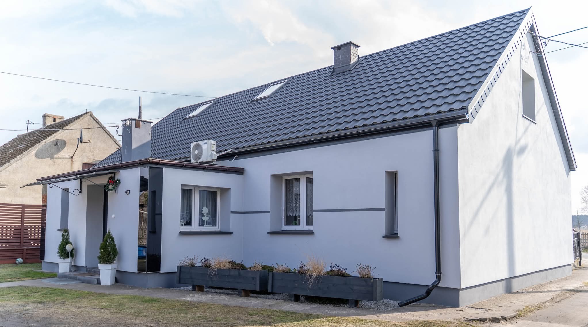 Biały dom jednorodzinny z szarą dachówką, po renowacji elewacji. Widoczne okna, klimatyzator i dekoracje roślinne. Ujęcie z poziomu ulicy w słoneczny dzień.