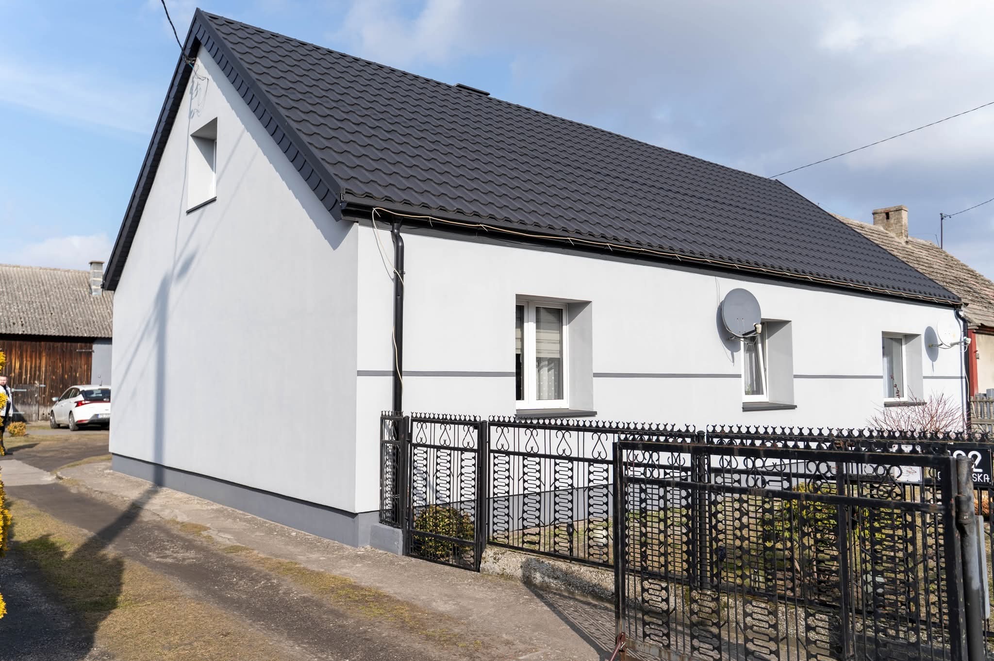 Biały dom jednorodzinny z ciemnym dachem, widoczny z boku, z czarnym ogrodzeniem i satelitami na elewacji. Świeżo pomalowana elewacja w spokojnej kolorystyce.