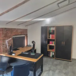 Biuro z biurkiem z drewnianym blatem, czarnymi nogami i krzesłami z niebieską tapicerką, regał na dokumenty z segregatorami, ściana z cegły i drewniane belki na suficie.