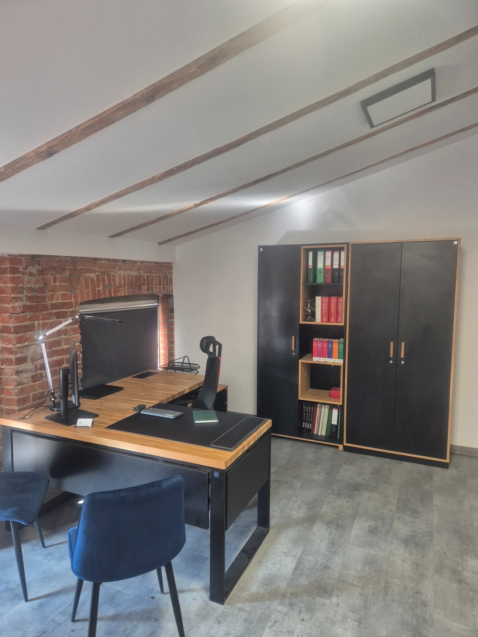 Biuro z biurkiem z drewnianym blatem, czarnymi nogami i krzesłami z niebieską tapicerką, regał na dokumenty z segregatorami, ściana z cegły i drewniane belki na suficie.