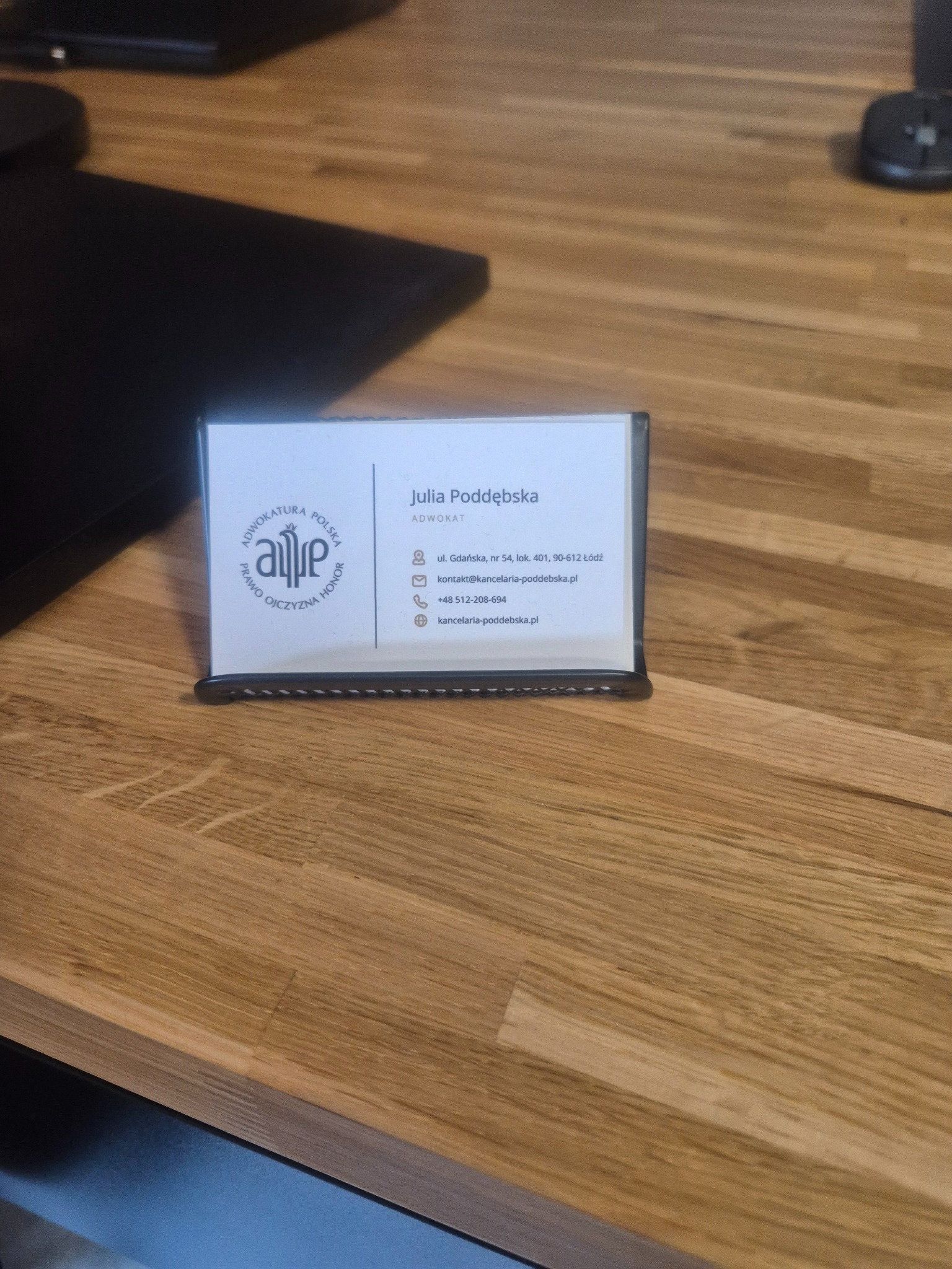 Wizytówka adwokata Julii Poddębskiej z logo, adresem i danymi kontaktowymi, umieszczona na drewnianym biurku.