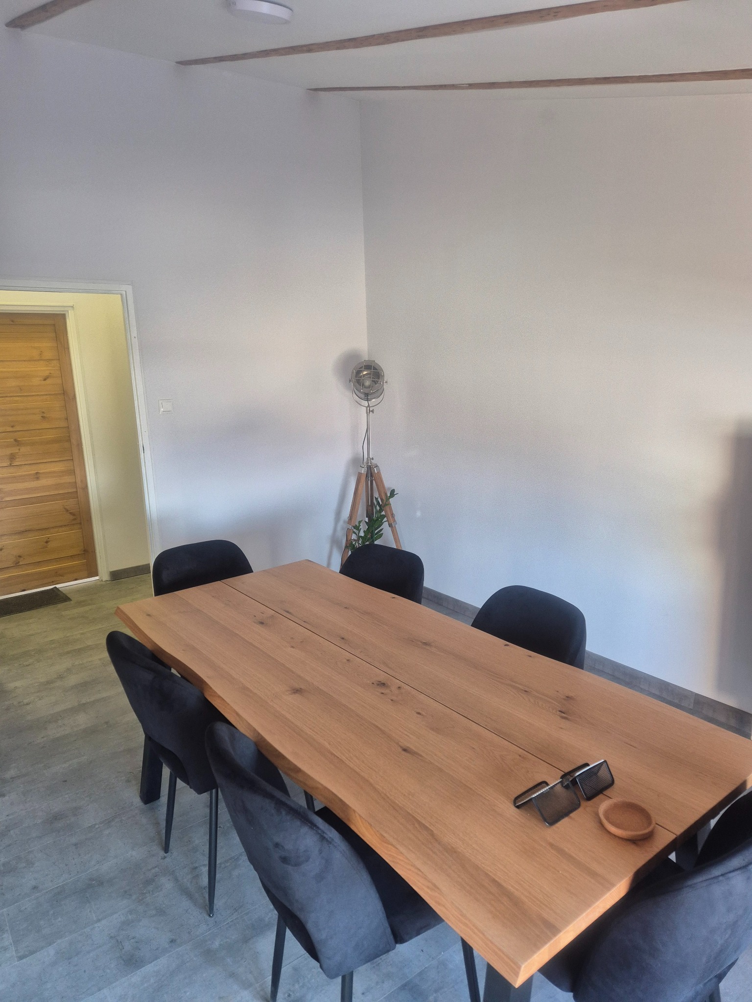 Wnętrze jadalni z dużym, drewnianym stołem, otoczonym przez sześć czarnych, welurowych krzeseł. Na stole leżą metalowe podkładki i mała, drewniana miseczka. W tle widoczna drewniana podłoga, białe...