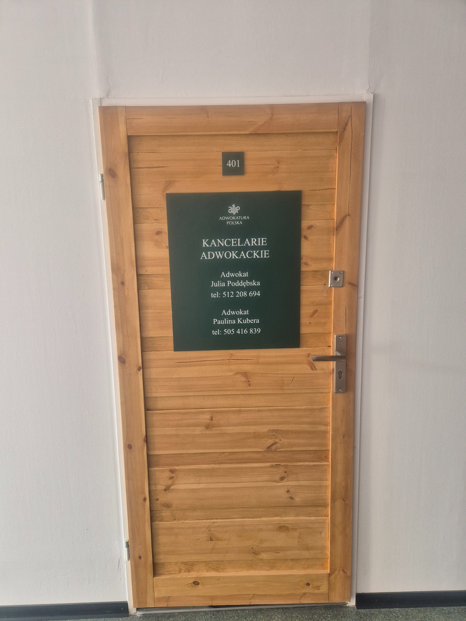 Drewniane drzwi do kancelarii adwokackiej z numerem 401 i tabliczką informacyjną z nazwiskami adwokatów Julii Poddębskiej i Pauliny Kubery oraz numerami telefonów.
