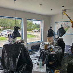 BART-BUD BARTOSZ ŁOŚ WYKOŃCZENIA WNĘTRZ - Prace przygotowawcze do gładzi: pomieszczenie zabezpieczone folią, narzędzia i materiały na stołach. Mężczyzna szlifuje sufit szlifierką teleskopową. Widok na ulicę przez okno.