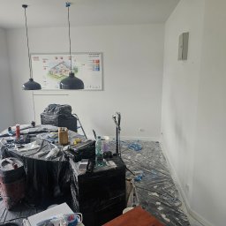 BART-BUD BARTOSZ ŁOŚ WYKOŃCZENIA WNĘTRZ - Białe ściany w trakcie malowania, zabezpieczone folią, widoczne narzędzia malarskie, lampy wiszące i plakat Ergo Hestia. Przygotowanie do malowania biura.