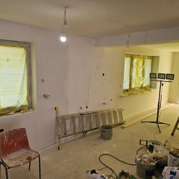 BART-BUD BARTOSZ ŁOŚ WYKOŃCZENIA WNĘTRZ - Wnętrze w trakcie szpachlowania ścian, okna zabezpieczone folią, widoczne narzędzia malarskie, drabiny i oświetlenie budowlane. Etap prac wykończeniowych.