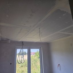 BART-BUD BARTOSZ ŁOŚ WYKOŃCZENIA WNĘTRZ - Montaż sufitu podwieszanego z płyty gipsowej w jasnym wnętrzu. Widoczne kable i elementy instalacji. Przez okno widać zieleń.