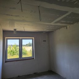 BART-BUD BARTOSZ ŁOŚ WYKOŃCZENIA WNĘTRZ - Surowy pokój po gładziowaniu ścian i sufitu, z widocznymi kablami i instalacją oświetleniową. Okno z widokiem na zieleń. Ściany przygotowane do dalszych prac wykończeniowych.