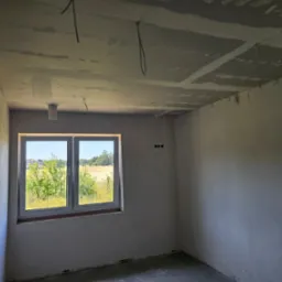 Surowy pokój po gładziowaniu ścian i sufitu, z widocznymi kablami i instalacją oświetleniową. Okno z widokiem na zieleń. Ściany przygotowane do dalszych prac wykończeniowych.