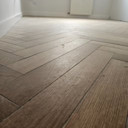 BART-BUD BARTOSZ ŁOŚ WYKOŃCZENIA WNĘTRZ - Parkiet ułożony w jodełkę w jasnym pomieszczeniu z białymi ścianami i grzejnikiem. Widoczne detale faktury drewna i precyzja ułożenia.