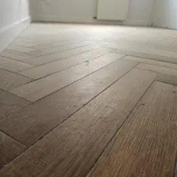 Parkiet ułożony w jodełkę w jasnym pomieszczeniu z białymi ścianami i grzejnikiem. Widoczne detale faktury drewna i precyzja ułożenia.