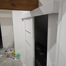 BART-BUD BARTOSZ ŁOŚ WYKOŃCZENIA WNĘTRZ - Białe, przesuwne drzwi z trzema czarnymi paskami, zamontowane w nowym, wykończonym wnętrzu. W tle widoczna drewniana belka, a na podłodze materiały budowlane.