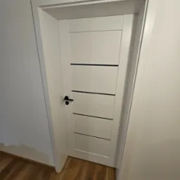 Nowoczesne, białe drzwi wewnętrzne z czarnymi, poziomymi akcentami i czarną klamką, wstawione w białą ościeżnicę, na tle ściany i drewnianej podłogi.