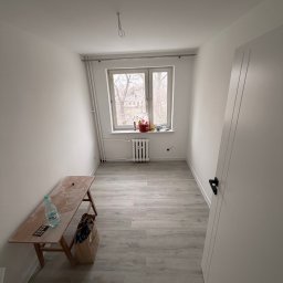 BART-BUD BARTOSZ ŁOŚ WYKOŃCZENIA WNĘTRZ - Wnętrze pokoju po malowaniu na biało, widoczne świeżo pomalowane ściany, okno z grzejnikiem i drewniana ławka z butelką wody. Podłoga wyłożona panelami.