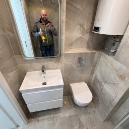 BART-BUD BARTOSZ ŁOŚ WYKOŃCZENIA WNĘTRZ - Wykończona łazienka z marmurowymi płytkami, białą armaturą i bojlerem. W lustrze odbija się wykonawca z narzędziami, prezentujący efekt końcowy.