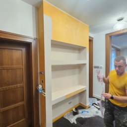 BART-BUD BARTOSZ ŁOŚ WYKOŃCZENIA WNĘTRZ - Wnęka z półkami po malowaniu, z żółtą górą i białymi półkami. Widoczny malarz z narzędziami i zabezpieczona podłoga. Ujęcie z perspektywy, z widocznym oświetleniem.