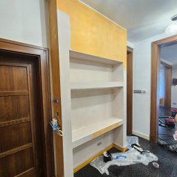 BART-BUD BARTOSZ ŁOŚ WYKOŃCZENIA WNĘTRZ - Wnęka z półkami w trakcie malowania na żółto, zabezpieczona folią podłoga, widoczne narzędzia malarskie i pędzel. Częściowo pomalowana ściana.